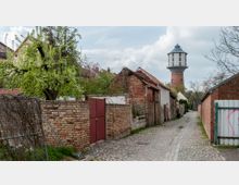 Schmale, gepflasterte Gasse mit Backsteinmauern und kleinen Gebäuden auf beiden Seiten, im Hintergrund ein historischer Wasserturm mit weißem und rotem Mauerwerk. Ein blühender Baum und ruhige ländliche Umgebung prägen die Szene.