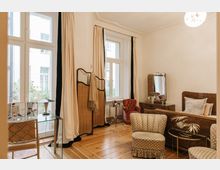 Heller, eleganter Wohnraum mit hellen Vorhängen an großen Fenstern, einem Holzboden, einer Polster-Sitzgruppe, einem Schminktisch mit Spiegel und einem historischen Charme. Im Raum stehen zusätzlich ein Raumteiler aus Holzgeflecht sowie ein roter Sessel und kleine Beistelltische.