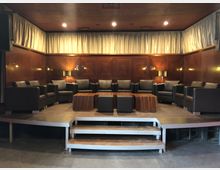 Ein kleiner Veranstaltungsraum mit einer erhöhten Bühne, auf der schwarze Ledersessel in einem Halbkreis um mehrere hölzerne Couchtische arrangiert sind. Der Raum hat holzvertäfelte Wände, cremefarbene Vorhänge und warme Beleuchtung.