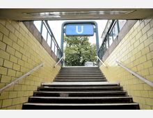 Treppenaufgang einer U-Bahn-Station mit gelben Fliesenwänden, Handläufen an beiden Seiten und einem blauen Schild mit weißem "U" über dem Ausgang. Im Hintergrund sind Bäume und ein Teil der städtischen Umgebung sichtbar.