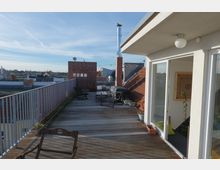 Große Dachterrasse mit Holzboden und Metallgeländer, ausgestattet mit Stühlen, einem Tisch und einem Grill. Im Hintergrund sind Backsteinmauern, Dächer und eine urbane Skyline unter klarem Himmel zu sehen.