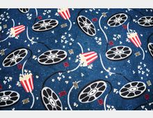 Blauer Teppich mit einem Muster aus schwarzen Filmrollen, verstreutem Popcorn und roten Popcornbechern mit weißen Streifen, ergänzt durch kleine Symbole in verschiedenen Farben. Die Gestaltung erinnert an ein Kino- oder Filmthema.