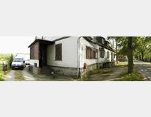 Weiß gestrichenes Gebäude mit Holzdach und Fensterläden, das an einer schmalen Straße liegt. Auf der linken Seite steht ein geparkter Lieferwagen; rechts sind Bäume und ein Gehweg sichtbar.