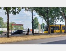 Straßenbahnhaltestelle "S Warschauer Straße" in Berlin mit gelber Straßenbahn im Hintergrund, umgeben von Bäumen und einer Straße im Vordergrund. Im Hintergrund sind Werbeplakate und parkende Fahrzeuge sichtbar.