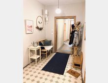 Ein gemütlicher Hausflur mit hellen Wänden, gemustertem Boden und Holztüren. Links steht eine kleine Sitzbank mit Kissen und einem Beistelltisch, rechts ein Holz-Kleiderständer mit Jacken und Hüten sowie eine Fußmatte mit der Aufschrift "hello".