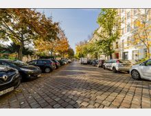 Straßenszene mit Kopfsteinpflaster, links und rechts geparkte Autos sowie Gebäuden mit weißer Fassade im Hintergrund; Bäume mit herbstlich gefärbtem Laub säumen die Straße.