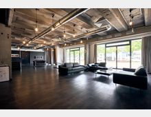 Industriell gestalteter Innenraum mit Sichtbetondecke, freiliegenden Lüftungsrohren und hängenden Glühbirnen. Der Raum ist mit schwarzen Ledersofas und Glastischen eingerichtet, großen Fenstern und Vorhängen, die Tageslicht hereinlassen.