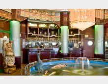 Innenraum eines asiatischen Restaurants mit einer Bar im Hintergrund, verziert mit Vasen und bunten Motiven an den Wänden. Im Vordergrund befindet sich ein kleiner Springbrunnen mit einer Entenstatue, daneben eine lachende Buddha-Statue aus Holz.