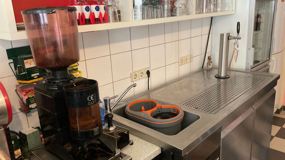Getränkezubereitungs- und Spülbereich mit Edelstahlarbeitsplatte, integrierter Spüle und Abtropffläche sowie Zapfhahn; daneben steht eine Kaffeemühle. Darüber befinden sich offene Regale mit Gläsern und Tassen vor einer weiß gefliesten Wand.