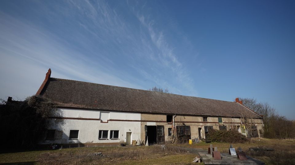 Langes, niedriges Wirtschafts- oder Scheunengebäude mit großem Satteldach und gemischter Fassade aus weiß verputzten und beige-grauen Bereichen, teils mit großen Toröffnungen und Fenstern. Davor liegt eine verwilderte Wiese mit einigen Holz- und Baumaterialresten, im Hintergrund stehen kahle Bäume unter blauem Himmel mit dünnen Wolkenschleiern.