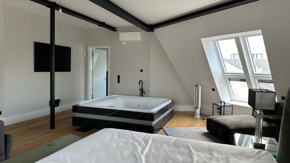 Modernes Dachgeschoss-Schlafzimmer mit sichtbaren schwarzen Stahlträgern und schrägen Wänden, in der Mitte mit einer großen Whirlpool-Badewanne auf dunklem Sockel. Rechts sorgen große Dachflächenfenster für Tageslicht; Holzboden, TV an der Wand und wenige Möbel ergänzen den Raum.