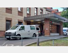 Außenansicht eines Krankenhauseingangs mit der Beschilderung "Notaufnahme" an einer Backsteinfassade. Vor dem Eingang stehen ein weißer Krankenwagen und ein weiteres Fahrzeug, der Bereich ist überdacht und von Fenstern sowie roten Akzenten geprägt.