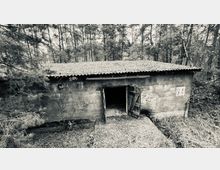 Schwarz-weiß-Fotografie eines kleinen, rechteckigen Gebäudes mit Wellblechdach, grober Betonfassade und einer offenen Holztür. Das Gebäude steht inmitten eines Waldgebietes, umgeben von Bäumen und dichtem Unterholz.