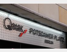 Schild eines Q-Park-Parkhauses am Potsdamer Platz in Berlin. Der Hintergrund besteht aus silbern glänzendem Riffelblech, mit schwarzer Schrift und roten Akzenten.