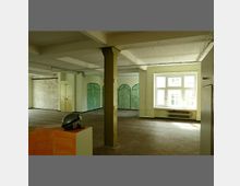 Ein leerer, offener Innenraum mit sichtbarer Stützsäule in der Mitte, großen Fenstern, die Tageslicht hereinlassen, sowie einem Parkettboden. An einer Wand befinden sich grüne, bogenförmige Türen, und daneben ist eine unverputzte Ziegelwand erkennbar.