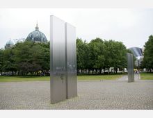 Denkmal mit mehreren hohen, vertikalen Metalltafeln auf einem gepflasterten Platz, umgeben von grünen Bäumen. Im Hintergrund ist die Kuppel des Berliner Doms sowie moderne Gebäudearchitektur sichtbar.
