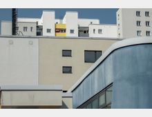 Moderner Gebäudekomplex mit weißen und beigen Fassaden sowie Fenstern in dunklen Rahmen, im Vordergrund ein geschwungener blauer Baukörper mit Glasfenstern. Im Hintergrund ist ein gelbes Balkondetail mit einem roten Sonnenschirm sichtbar.