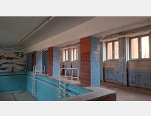 Verlassenes Hallenbad mit leerem, blau gefliestem Becken und mehreren Metallleitern entlang des Beckenrands. Blaue und rotbraune Fliesenpfeiler tragen die Decke; an der rechten Wand befinden sich Fenster, freiliegende Rohrleitungen und abblätternde Fliesen.