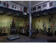 Innenraum eines Puppenmuseums mit gelb gestrichenen Wänden, Glasschränken und Vitrinen, die zahlreiche Marionetten und Puppen zeigen. Oberhalb gibt es eine Galerie mit blauem Geländer, an der weitere Exponate und Gemälde ausgestellt sind.