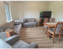 Helles Wohn- und Esszimmer mit zwei grauen Sofas, Holzboden und großem Fenster an der linken Wand. Rechts stehen ein runder Holzesstisch mit gepolsterten Stühlen sowie ein Fernseher auf einem Eckschrank.