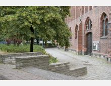 Backsteinfassade einer Kirche mit gotischen Spitzbogenfenstern und einer verzierten Eingangstür aus Metall; davor ein gepflasterter Weg, umgeben von Bäumen und Grünflächen. Im Vordergrund sind niedrige Mauern und Gehwege aus Stein zu sehen.