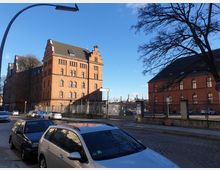 Straßenszene mit historischen Backsteingebäuden im Hintergrund, darunter ein markantes Gebäude mit Treppengiebeln. Im Vordergrund sind geparkte Autos auf einer Kopfsteinpflasterstraße zu sehen, flankiert von einem hohen Metallzaun. Ein kahler Baum und eine Straßenlaterne rahmen das Bild links und rechts ein.