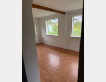 Leerer Innenraum mit hellem Laminatboden, weißen Wänden und freiliegendem Holzbalken unter der Decke. Zwei große Fenster lassen Tageslicht herein und bieten Blick ins Grüne sowie auf Nachbarhäuser.
