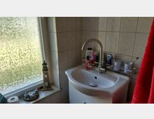 Waschbecken in einem gefliesten Badezimmer mit hohem Wasserhahn und Ablage mit Pflegeartikeln. Links befindet sich ein Fenster mit strukturierter Milchglasscheibe und Fensterbank mit Dekoration.