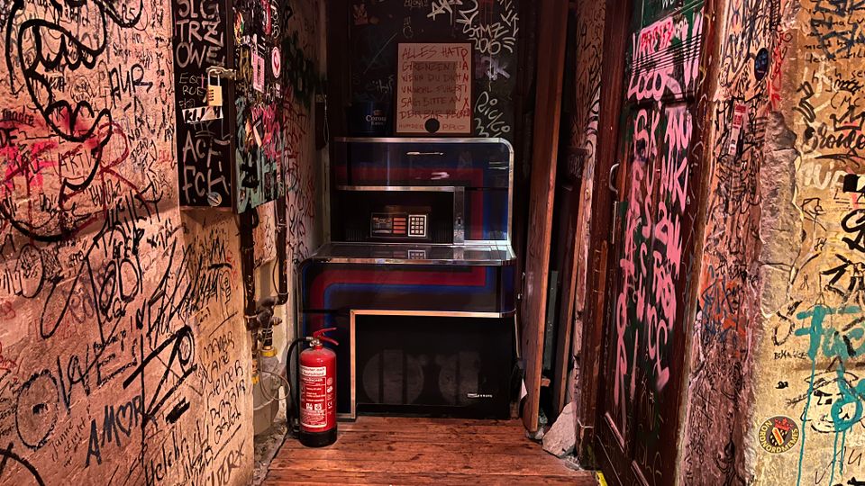 Schmaler Innenflur mit Holzboden, dessen Wände und Türen dicht mit Graffiti und Aufklebern bedeckt sind. Am Ende des Gangs stehen ein beleuchteter Automat bzw. eine Jukebox mit Tastaturfeld sowie ein Feuerlöscher und eine niedrige Box auf dem Boden.