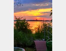 Sonnenuntergang an einem See mit orange-gelb leuchtendem Himmel und reflektierendem Wasser, umgeben von Schilf und Gräsern. Im Vordergrund ein kleiner Steg mit Metallgeländer und einer grünen Gießkanne.
