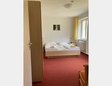 Ein einfach eingerichtetes Schlafzimmer mit einem Doppelbett auf rotem Teppichboden, weißer Bettwäsche und gefalteten Handtüchern. Im Hintergrund befinden sich ein Fenster mit gelben Vorhängen, ein Heizkörper und ein kleines Wandbild.