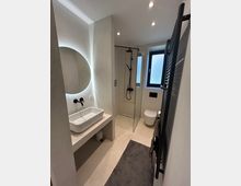 Modernes Badezimmer mit minimalistischer Gestaltung, einem runden beleuchteten Spiegel über einem rechteckigen Waschbecken, einer bodenebenen Dusche mit Glaswand, einer Wandtoilette und einem dunklen Handtuchheizkörper. Der Raum ist in hellen Tönen gehalten und verfügt über ein Milchglasfenster, das diffuses Licht einlässt.