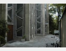 Außenansicht eines modernen Betongebäudes mit geometrischen Fenstern, die diagonal unterteilt sind. Im Vordergrund ein gepflasterter Hof mit einer Fahrradlampe und einer Mauer, umgeben von Bäumen im Hintergrund.