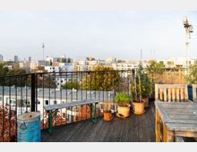 Dachterrasse mit Holzboden, Geländer aus schwarzem Metall und mehreren Topfpflanzen, darunter Kräuter und kleine Sträucher. Im Hintergrund Blick auf städtische Gebäude, die Berliner Fernsehturm und einen Kran.