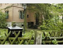 Backsteingebäude mit grünen Fensterläden, umgeben von dichtem Gebüsch und Bäumen. Im Vordergrund stehen ein Zaun aus Holz und ein schwarzer Briefkasten, daneben eine Straßenlaterne.