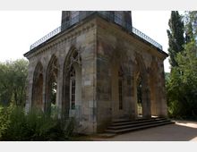 Historisches Gebäude mit neugotischer Architektur, gekennzeichnet durch spitzbogige Arkaden und verzierte Fensterrahmen. Das Bauwerk ist von Bäumen und Grünflächen umgeben, mit einer kleinen Treppe, die zu den Eingängen führt.