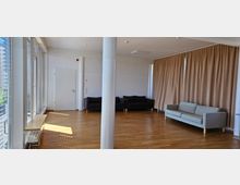 Ein heller Raum mit Parkettboden, weißen Wänden, großen Fenstern mit Jalousien und beigefarbenen Vorhängen. Im Hintergrund stehen mehrere Sofas, eine helle Couch und zwei dunkle Couches, sowie eine geschlossene Tür an der Wand.