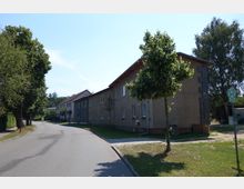 Wohnstraße mit einem zweistöckigen Gebäude aus Putz und Ziegeln auf der rechten Seite, umgeben von Bäumen und Grünflächen. Im Vordergrund ein Gehweg, vereinzelt gepflanzte junge Bäume und ein Haltestellenschild auf einer kleinen Wiese.