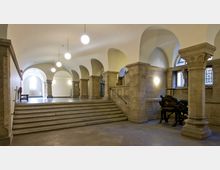 Historischer Innenraum mit gewölbten Decken, massiven Säulen und einer Treppe aus Stein. An der rechten Seite steht eine antike Maschine, und durch Buntglasfenster fällt Licht in den Raum.