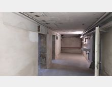 Ein einfacher, karger Kellerraum mit weiß verputzten Wänden, Betonboden und einem Waschbecken an der rechten Wand. An der Decke verlaufen Rohre und Kabel, und die Beleuchtung ist spärlich.