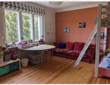 Helles Kinderzimmer mit breitem Fensterband und gestreiften Raffrollos, Holzdielenboden und Blick ins Grüne. Im Raum stehen ein runder Tisch mit Hockern und Spielzeugkisten, ein rotes Sofa vor einer orangefarbenen Akzentwand sowie ein Hochbett mit Leiter.
