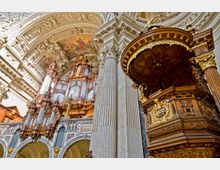 Prunkvoller Innenraum einer Barockkirche mit detaillierten goldenen und hölzernen Schnitzereien. Im Vordergrund eine reich verzierte Kanzel, dahinter eine große Orgel mit Pfeifen und ornatierten Verzierungen, flankiert von Säulen und kunstvoll gestalteten Gewölben.