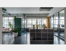 Modernes Büro mit großen Fenstern, die viel Tageslicht hereinlassen. Im Vordergrund steht ein quadratischer Tresen mit Pflanzen, links eine Sitzecke mit Sofa und rechts verglaste Besprechungsräume.