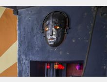 Eine metallische Maske mit leuchtenden Augen ist an einer dunkelblauen, strukturierten Wand angebracht. Darunter befindet sich eine Nische mit rötlich-violett beleuchteten Elementen.