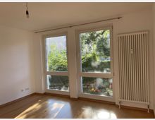 Ein heller, leerer Raum mit Holzfußboden, weißen Wänden und großen bodentiefen Fenstern, die auf einen begrünten Außenbereich blicken. An der rechten Wand befindet sich ein weißer Heizkörper, und an der Decke hängt eine einzelne Glühbirne.