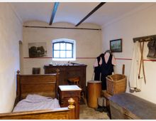 Kleines historisches Schlafzimmer mit einem Holzbett, Nachttisch und Kommode. An der Wand hängen Bilder, daneben befinden sich ein Wäschekorb, Kleidungsstücke und Alltagsgegenstände; das Mauerwerk und die Einrichtung wirken schlicht und rustikal.