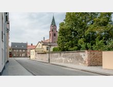 Straßenansicht einer Wohngegend mit gepflastertem Gehweg, flankiert von Gebäuden mit roten und grauen Dächern. Im Hintergrund ist eine hohe Backsteinkirche mit spitzem Turm und Uhr zu sehen, umgeben von großen Bäumen.