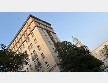 Schräg aufgenommene Ansicht eines hohen, rechteckigen Wohn- oder Bürogebäudes mit hellem Kachelmuster und vielen Fenstern, teils mit kleinen Balkonen. Im Vordergrund stehen große Bäume; rechts ragt hinter ihnen ein Turm mit grüner Kuppel vor blauem Himmel hervor.