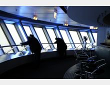 Innenansicht eines Aussichtsraumes mit großen, geneigten Panoramafenstern, durch die diffuses Licht fällt. Im Vordergrund sind Metallbarhocker und eine geschwungene Theke zu sehen, während Personen an der Brüstung stehen und hinausblicken.