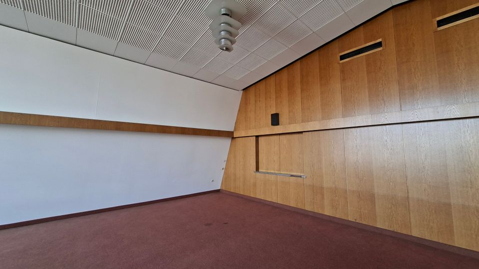 Leerer Innenraum mit rotem Teppichboden, weißer Wandfläche und großer Holzvertäfelung an der rechten Seite mit eingelassenen Lüftungsöffnungen und einer schmalen Ablage. An der Decke befinden sich perforierte Akustikplatten und eine zentrale Deckenleuchte.